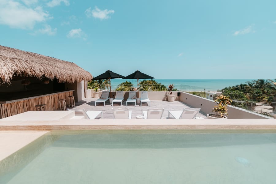 Ein Hotelpool mit Liegen unter einem Dach aus Palmstroh, dahinter das Meer