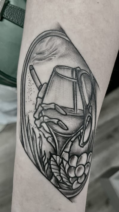 A unique black and grey illustrative tattoo in an oval frame. It depicts a skeleton hand holding a smoking cigarette and a glass of what appears to be whiskey or a similar drink. A pair of glasses hangs off the glass, and floral elements adorn the bottom.