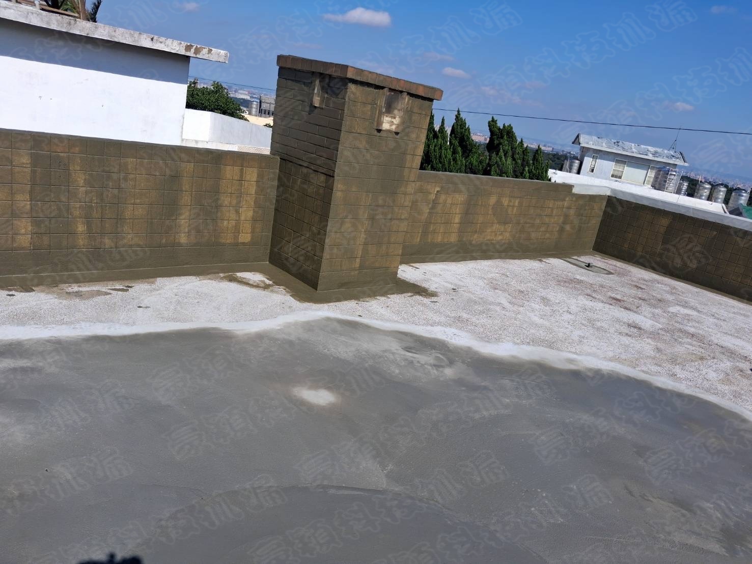 圖片展示了一個屋頂防水層的施工現場。屋頂表面塗覆了一層灰色的防水材料，部分區域已經完成塗覆，而其他區域仍待施工。牆面為磚砌結構，已出現污漬和發霉跡象，表明可能存在漏水問題。圖片在藍天白雲的背景下拍攝，清晰展示了施工現場的狀況。這個場景在漏水檢測和防水工程中具有重要意義，突出了防水層施工前的準備和必要性。