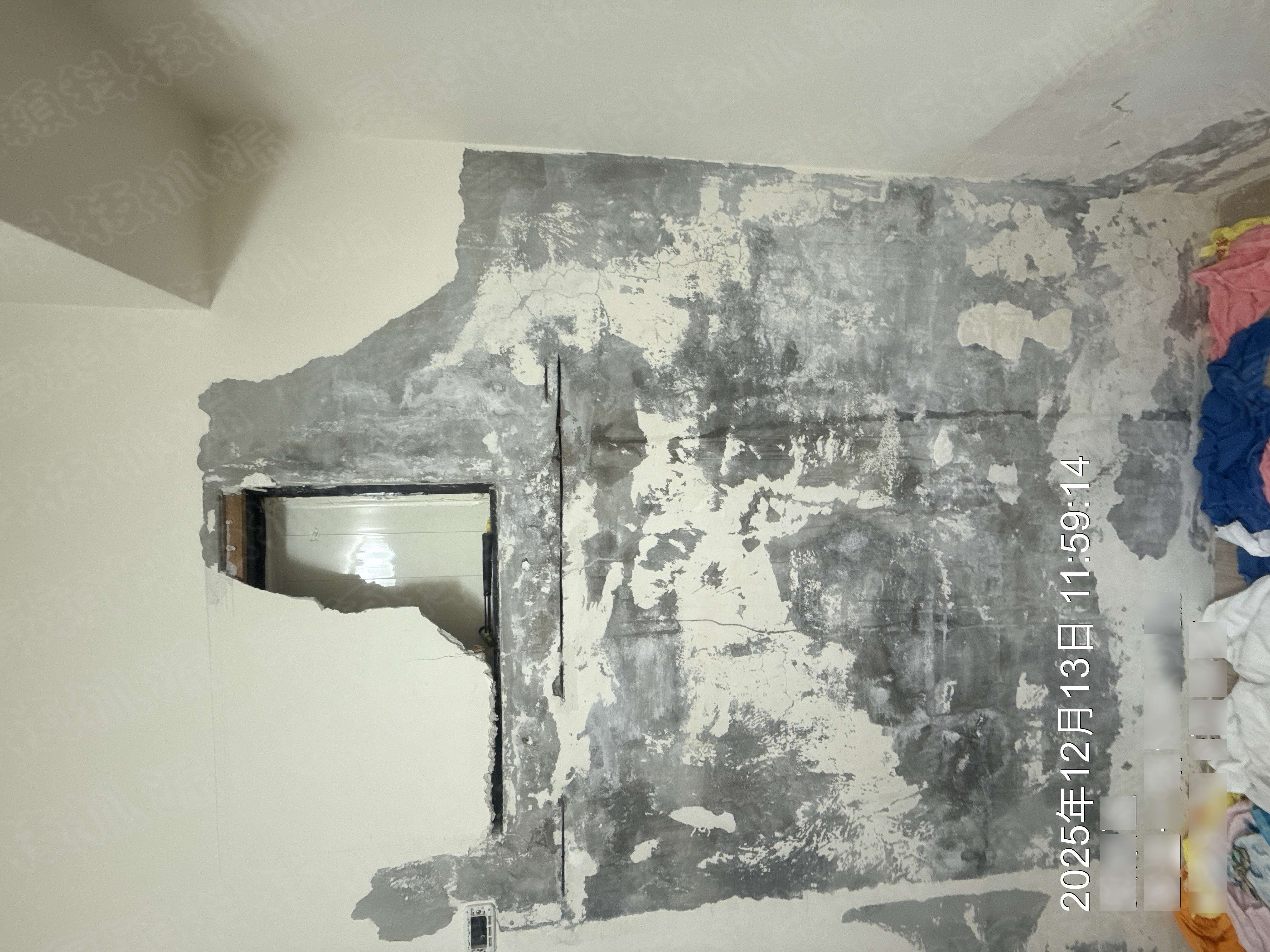 圖片展示了一面牆壁的剝落和壁癌問題。牆面出現大面積灰白色剝落，局部可見水泥基材，牆面下方有明顯的垂直裂縫。右側可見部分堆放的物品。圖片為現場實拍，記錄了牆面問題的真實狀態。