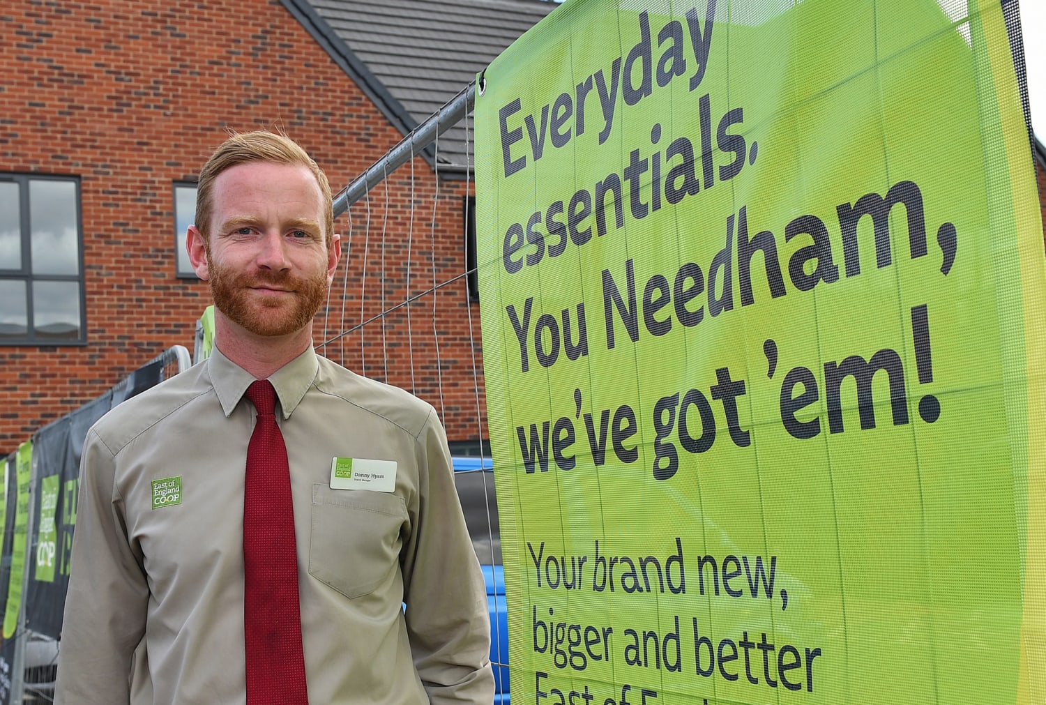 The manager of the new Needham Market food store is standing on-site, next to signage on fencing that reads, “Everyday essentials. You Needham? We’ve got ’em!”