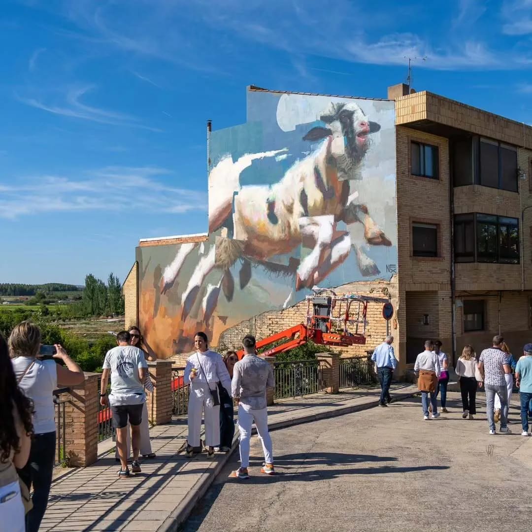 Un mural de una cabra blanca en un edificio, con personas observando en la calle.