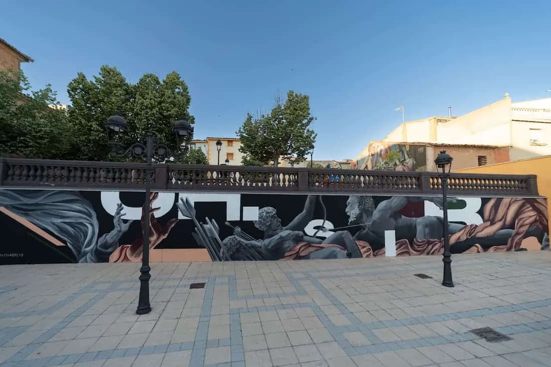 Mural urbano con figuras humanas en una pared, con una barandilla y farolas en primer plano.