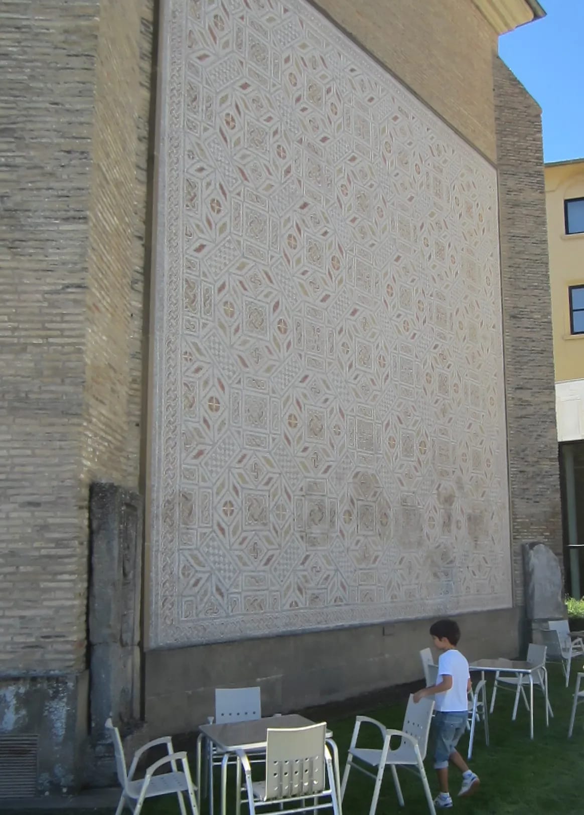 Un niño pasa por delante de una pared grande con un diseño geométrico en un patio con mesas y sillas.
