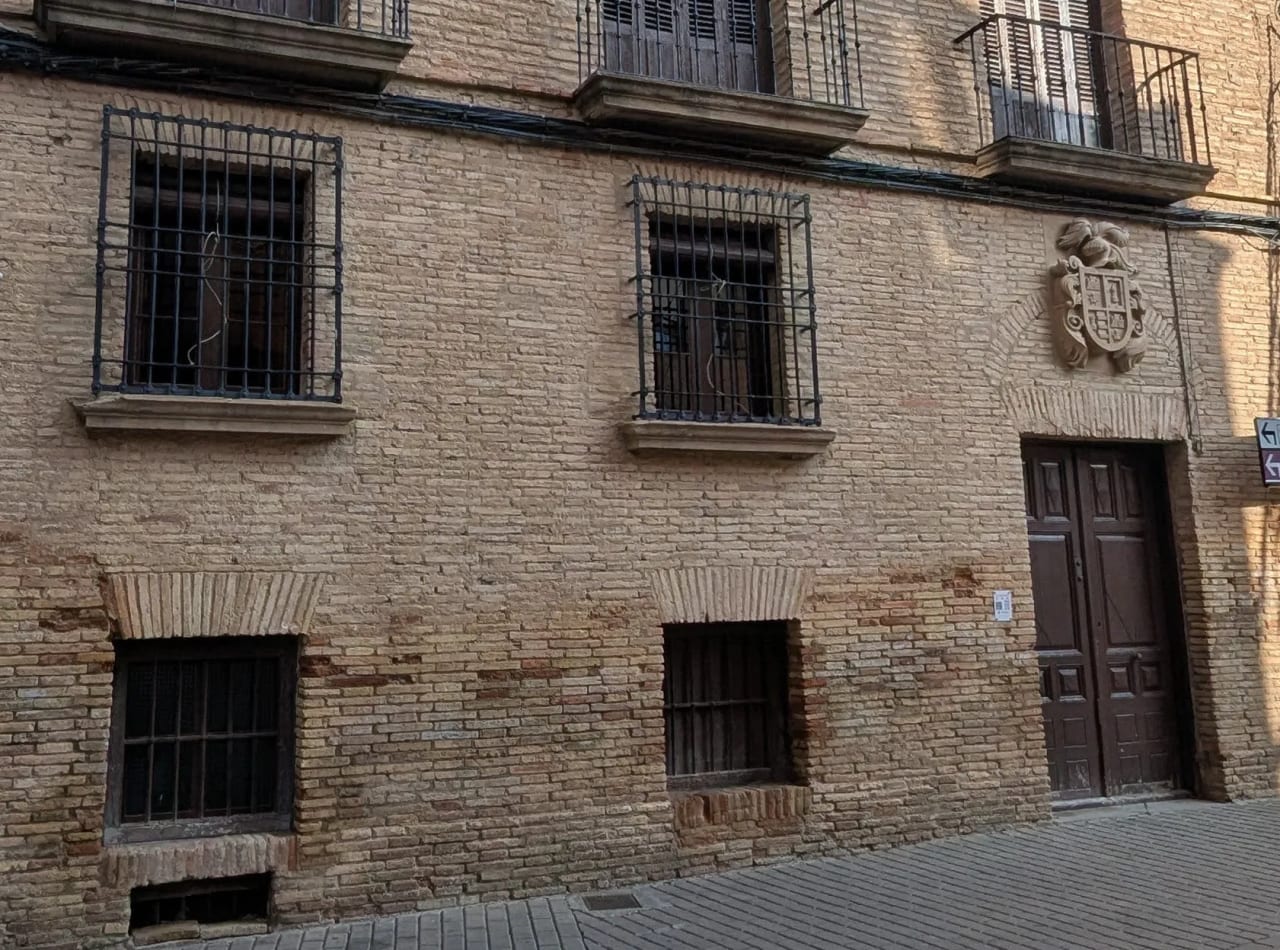 Fachada de ladrillo con ventanas enrejadas y puerta de madera con emblema.