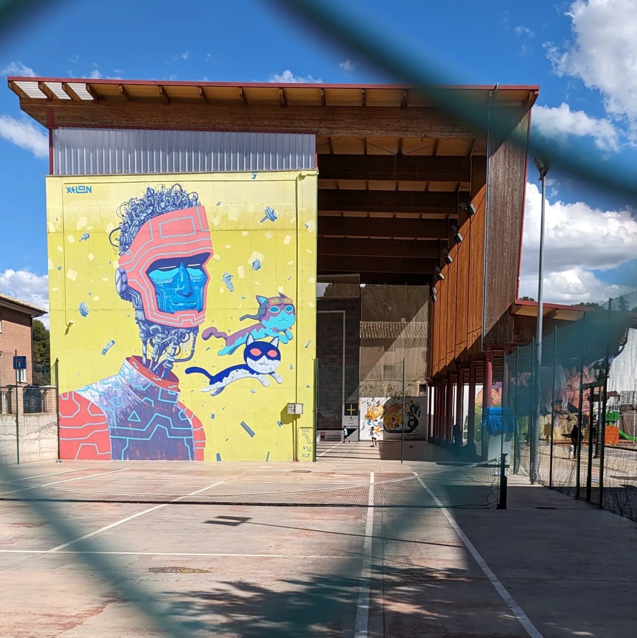 Mural de un hombre con gafas y gatos volando en una pared amarilla de un edificio de madera con pistas de tenis.