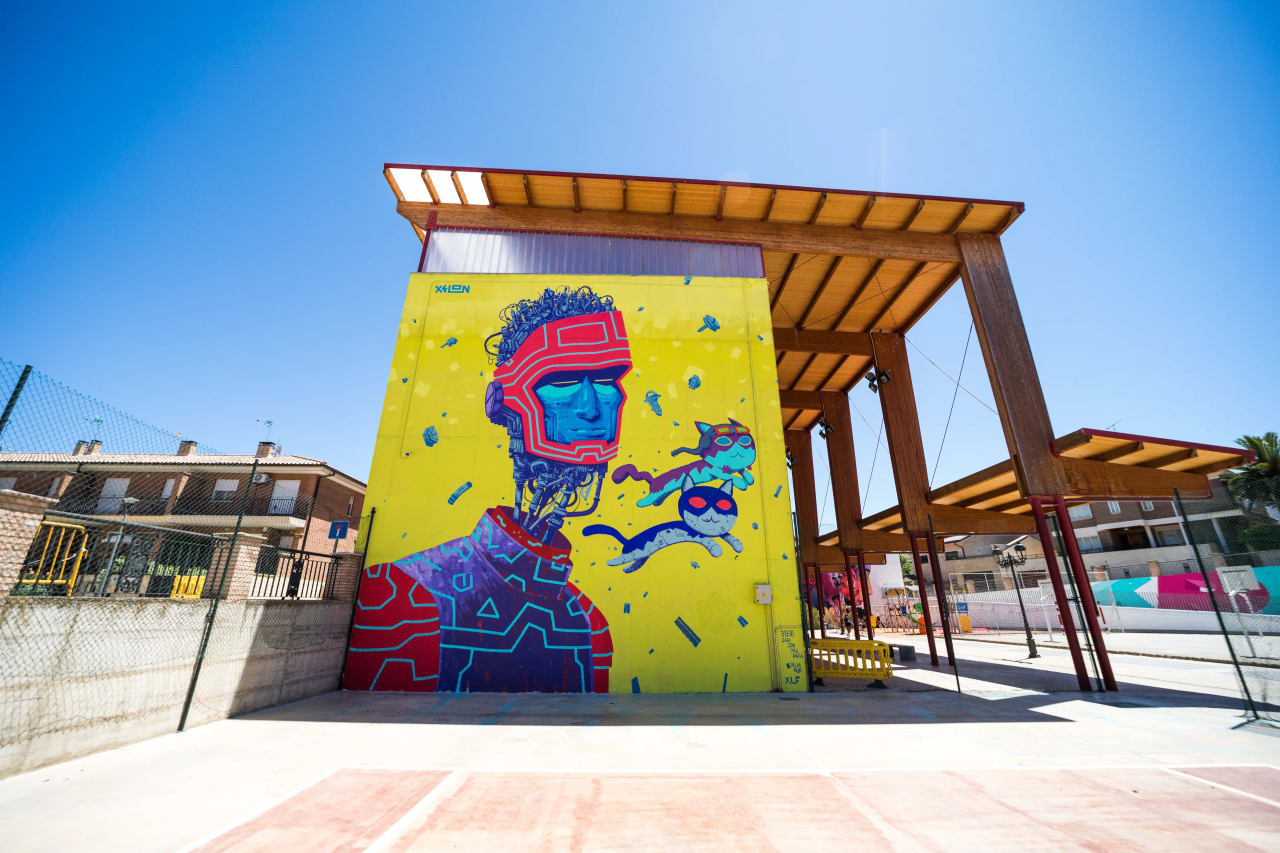 Mural de un hombre con gafas de realidad virtual y dos gatos voladores en una pared amarilla.