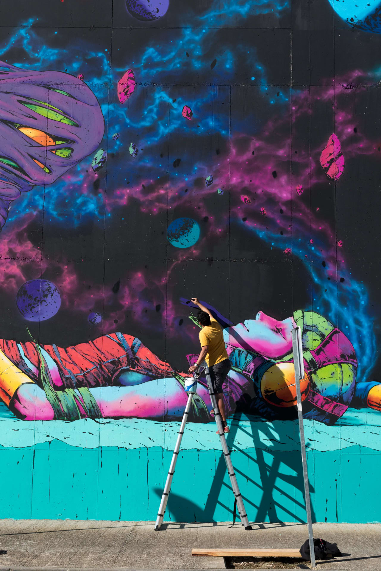 Un artista pinta un mural vibrante en una pared exterior mientras está subido a una escalera.