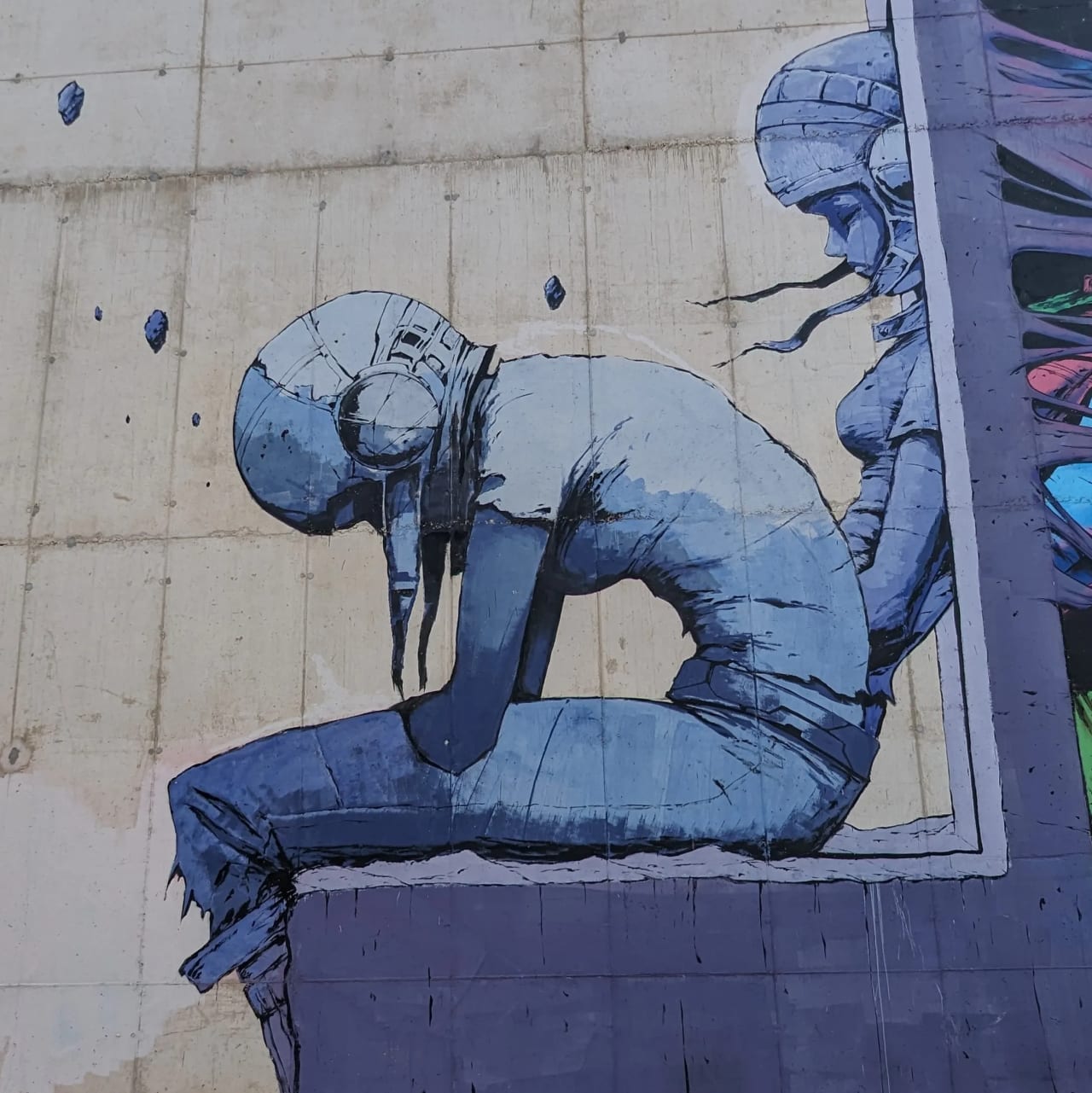 Mural de una persona sentada con la cabeza gacha en tonos azules sobre pared de cemento.