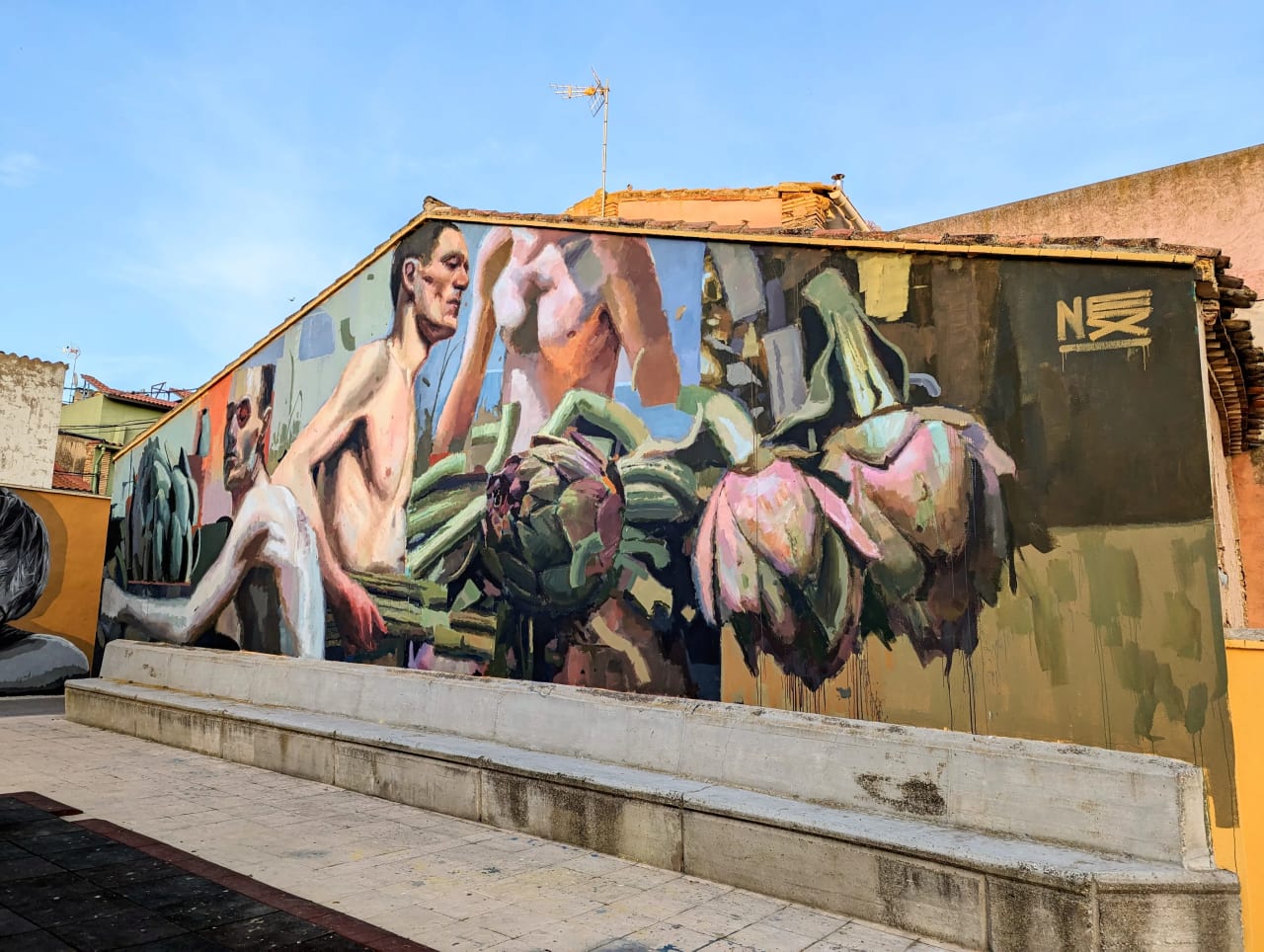 Mural de dos hombres desnudos con flores en un fondo verde en una pared exterior.