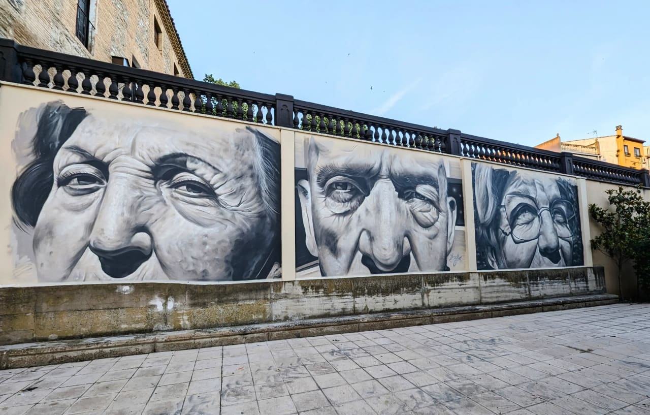 Mural con tres retratos realistas en blanco y negro de rostros de personas mayores en una pared de piedra.