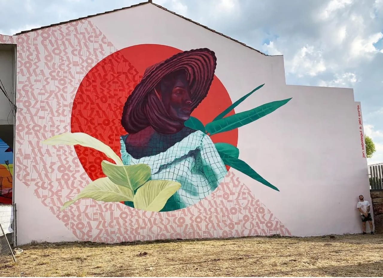 Mural de una mujer con sombrero en una pared con fondo rojo y texto rosa.
