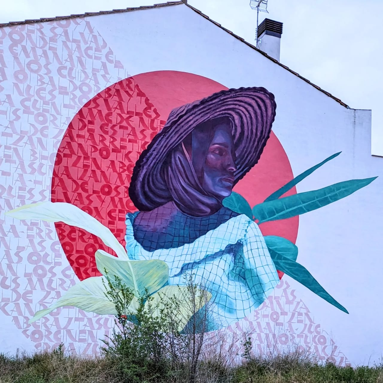 Mural de una mujer con sombrero y hojas verdes en una pared blanca.