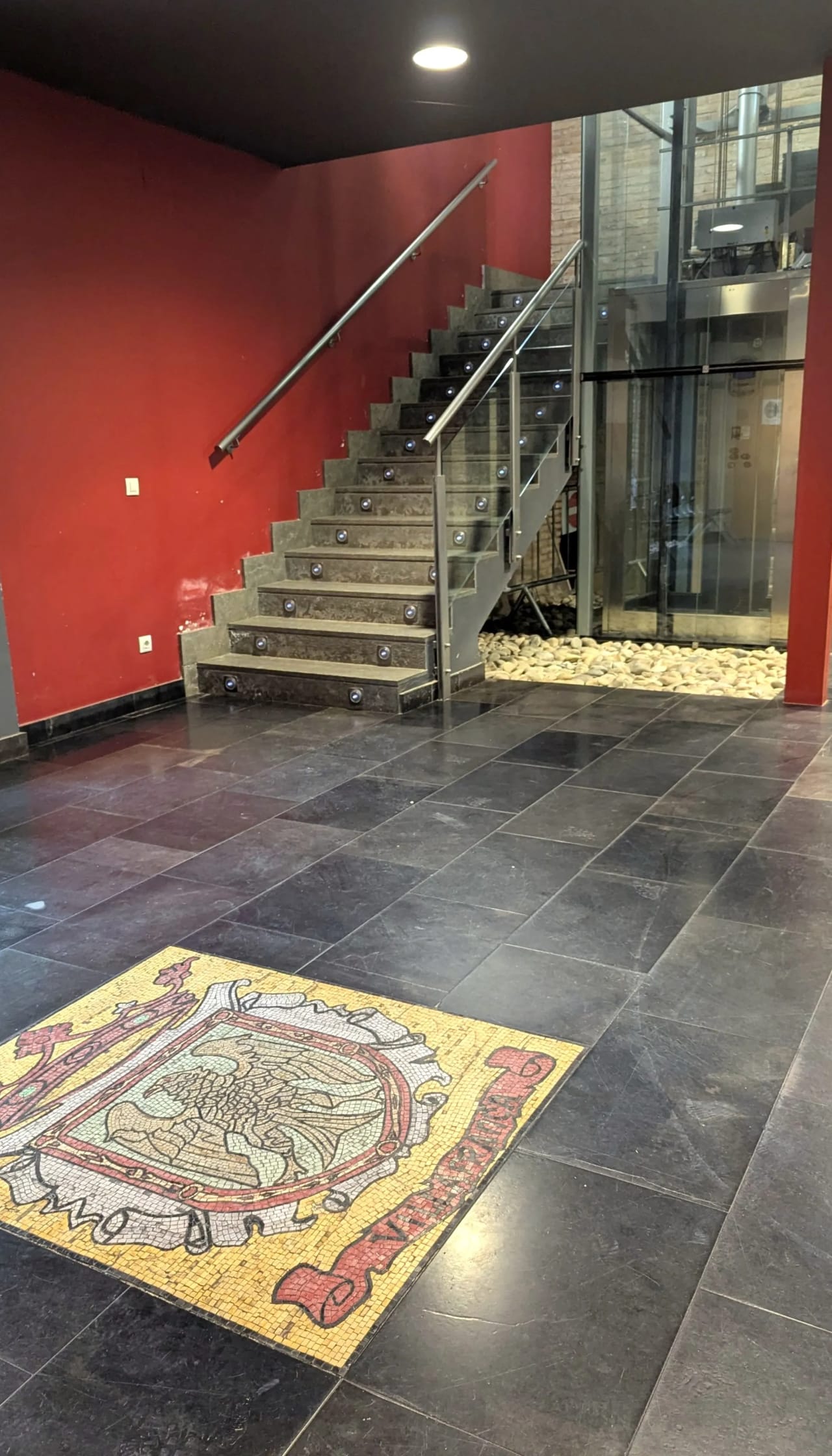 Escalera de metal con barandilla junto a pared roja y suelo de baldosas con diseño mosaico.