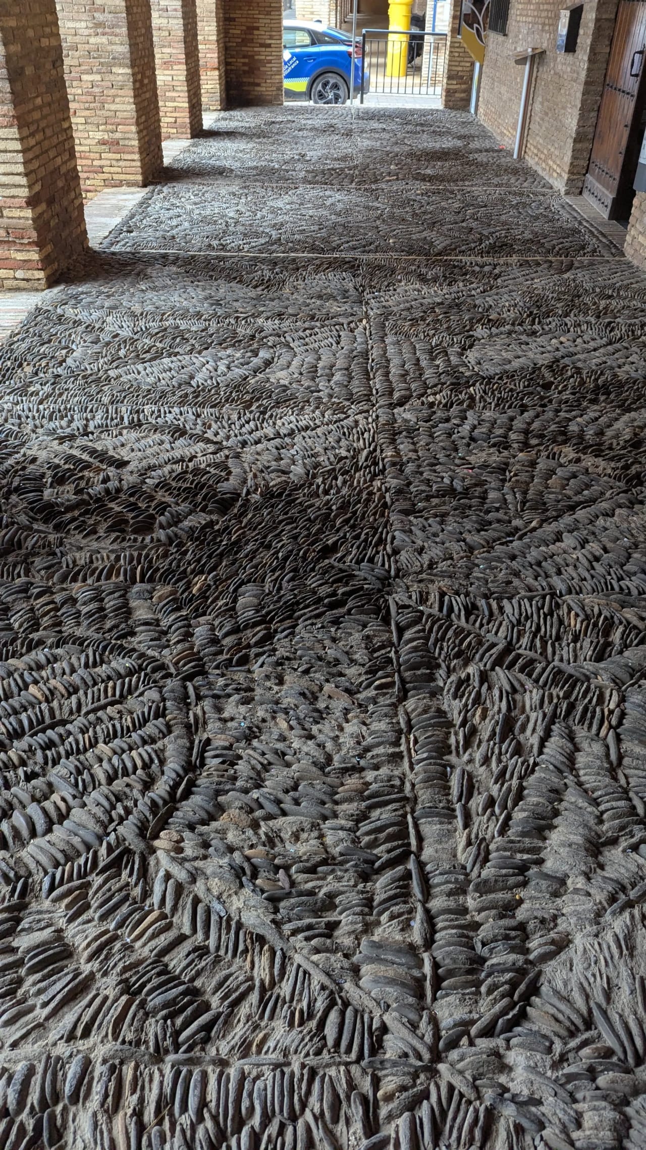 Pavimento de conchas en un pasillo con columnas de ladrillo y un coche azul al fondo.