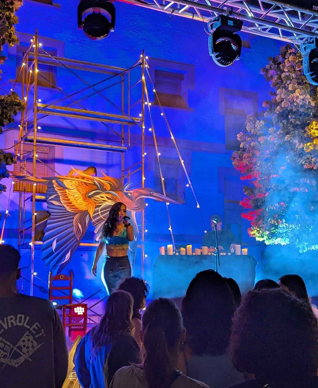 Mujer canta en un escenario con iluminación azul y una gran ave decorativa detrás de ella.