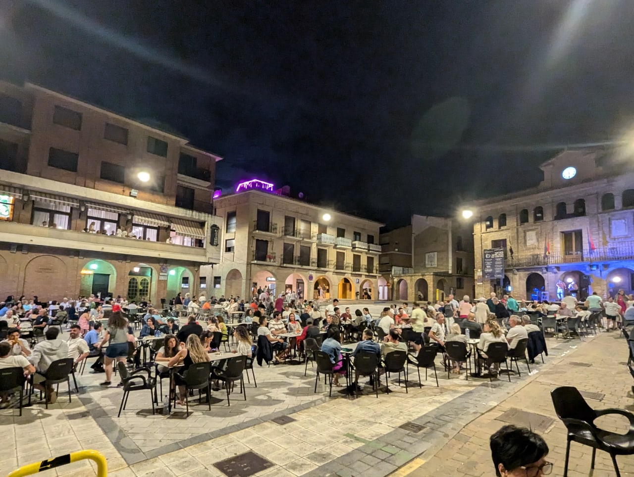 Plaza llena de gente sentada en mesas y sillas en una noche oscura.
