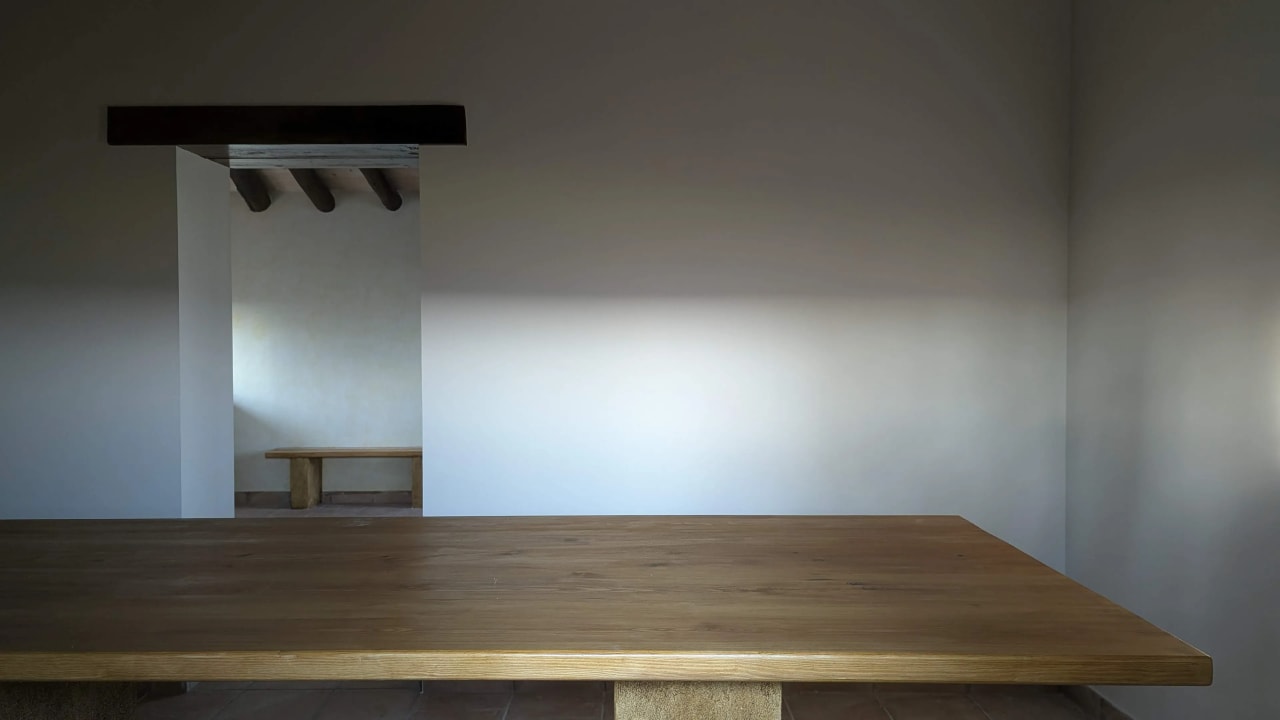 Mesa de madera en una habitación con paredes blancas y una abertura en el fondo con otro banco.