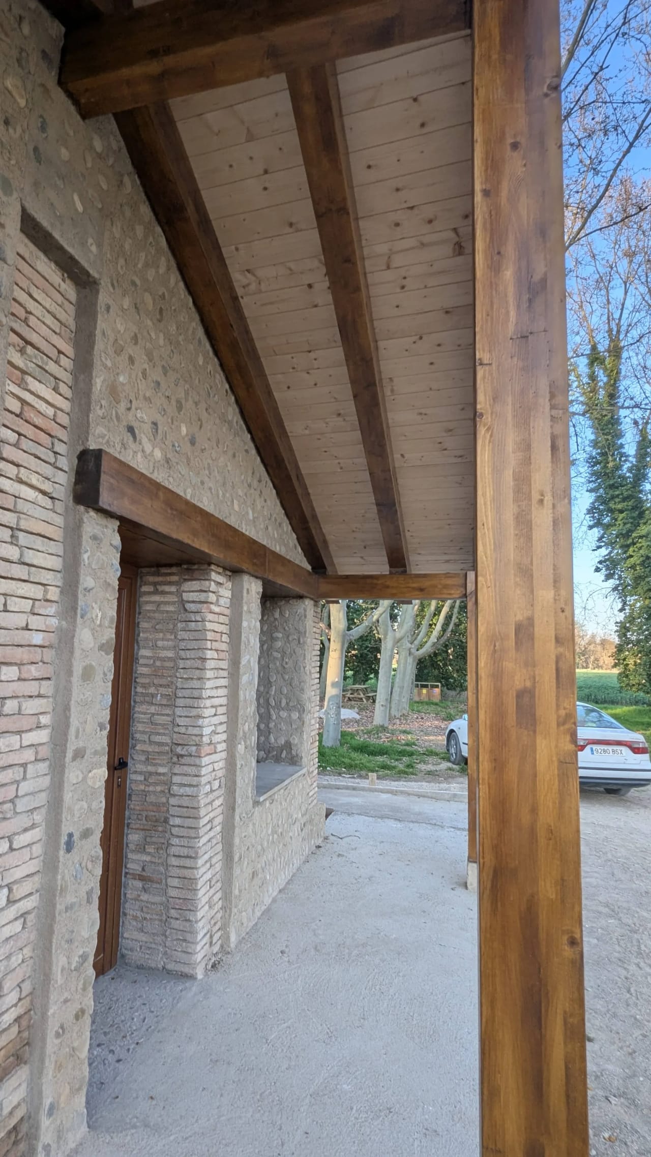 Pórtico de madera con vigas oscuras y techo de madera clara sobre base de piedra y ladrillo.