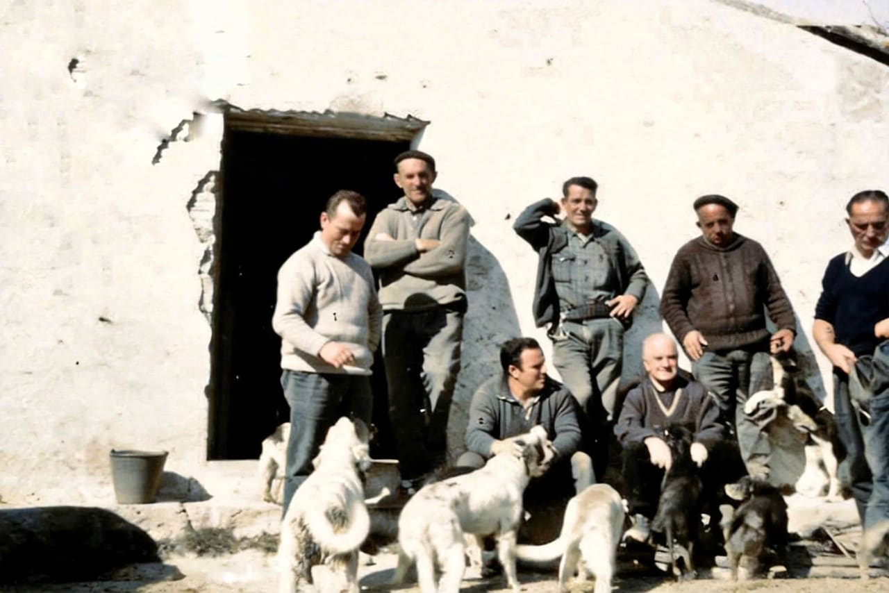 Siete hombres posan con varios perros delante de una casa blanca con la puerta abierta.