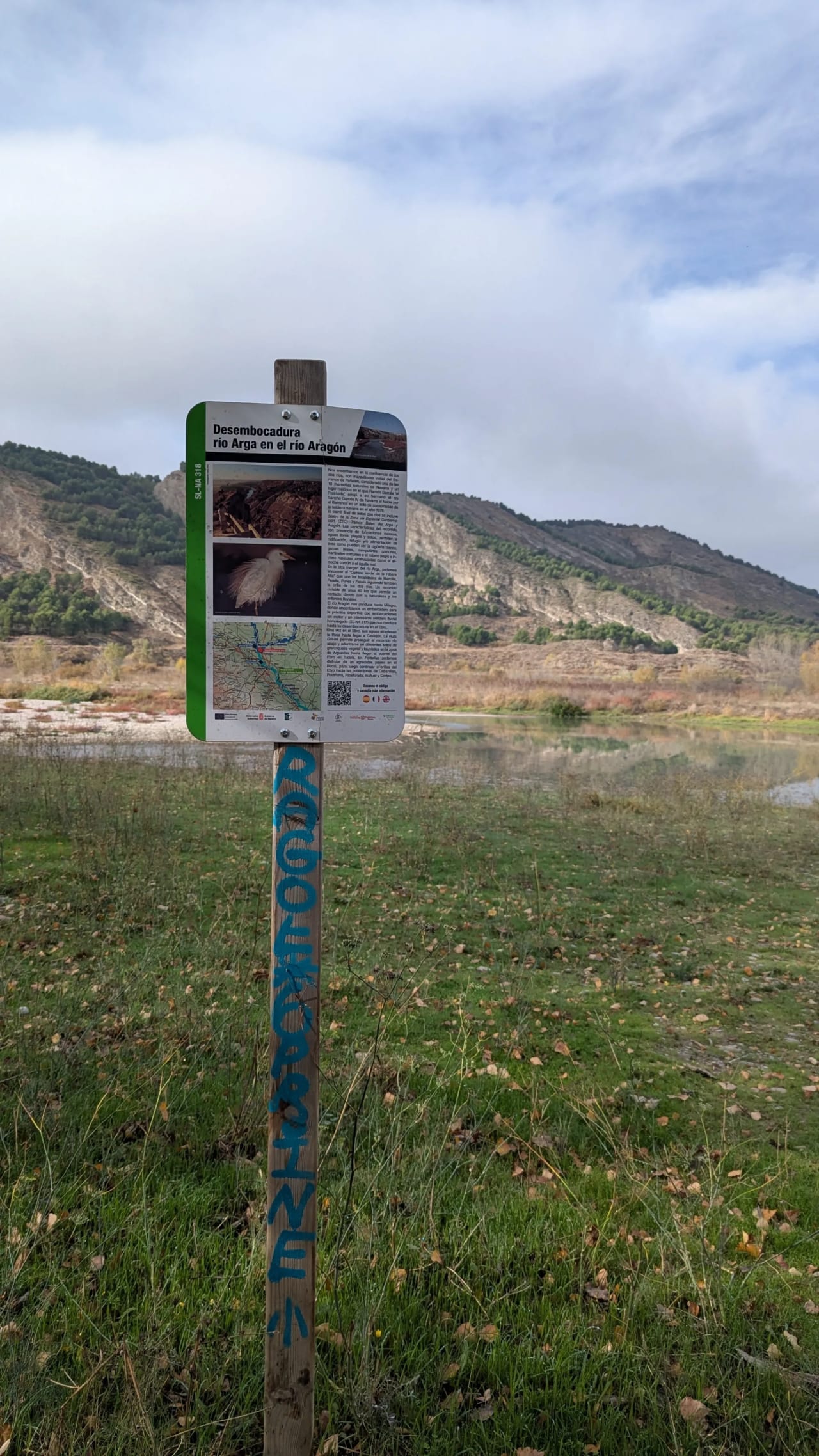 Señal informativa en un poste de madera en un paisaje natural con un río y montañas al fondo.