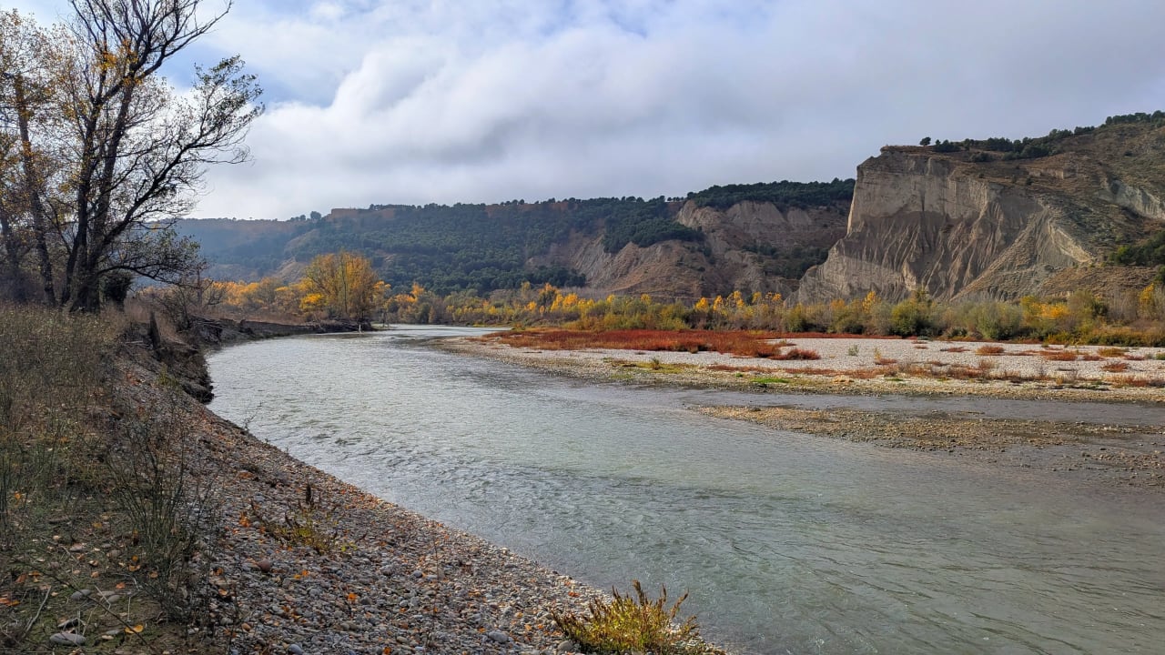 Río serpenteante en un paisaje montañoso con árboles de otoño y nubes.
