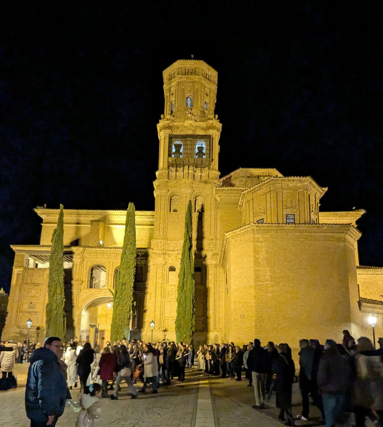 Fachada iluminada de la Parroquia de Santa Eufemia con su torre y multitud de personas reunidas afuera.