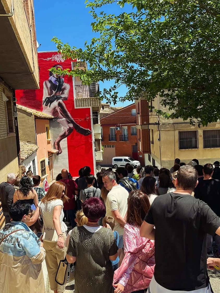 Multitud reunida en una calle frente a un mural grande y rojo de un hombre desnudo.