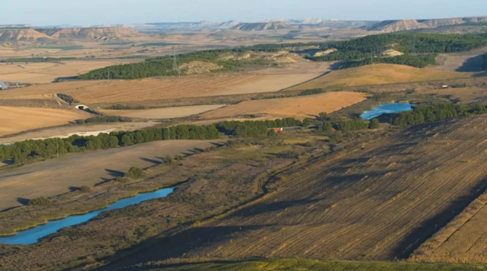 Paisaje rural con campos de cultivo, arroyos y montañas al fondo.