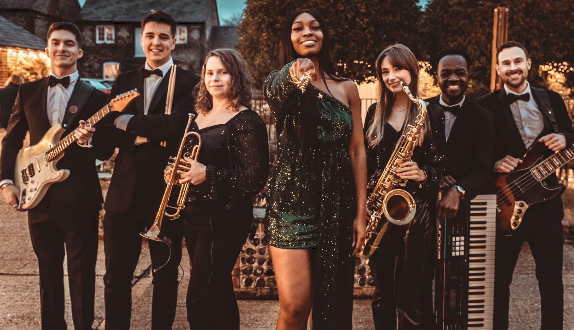 Dancefloor Magic: Seven band members posing with guitars, keyboard, trumpet and saxophone outdoors