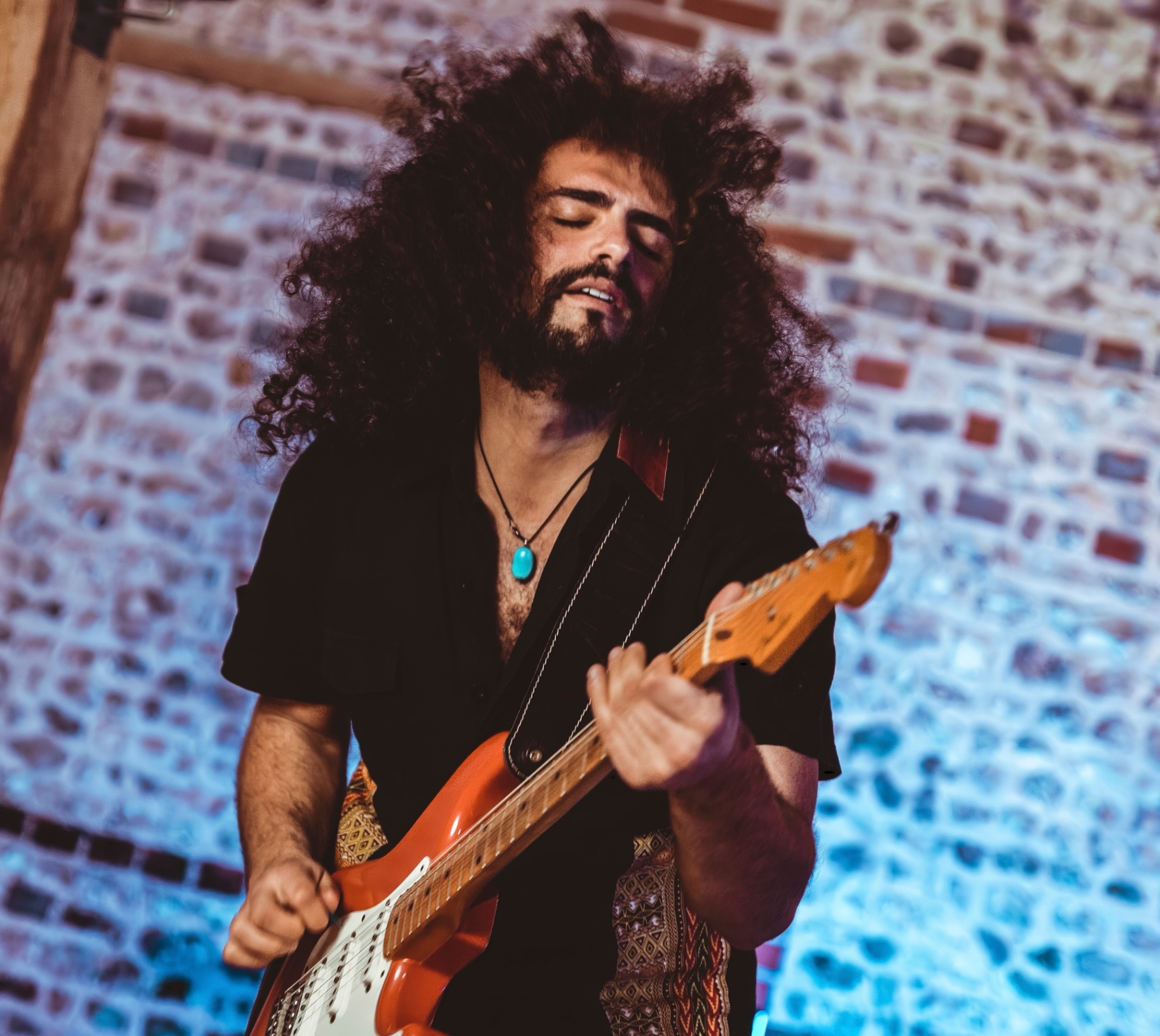 On The Rocks: Guitarist with long curly hair playing an orange electric guitar against a blue-lit brick wall