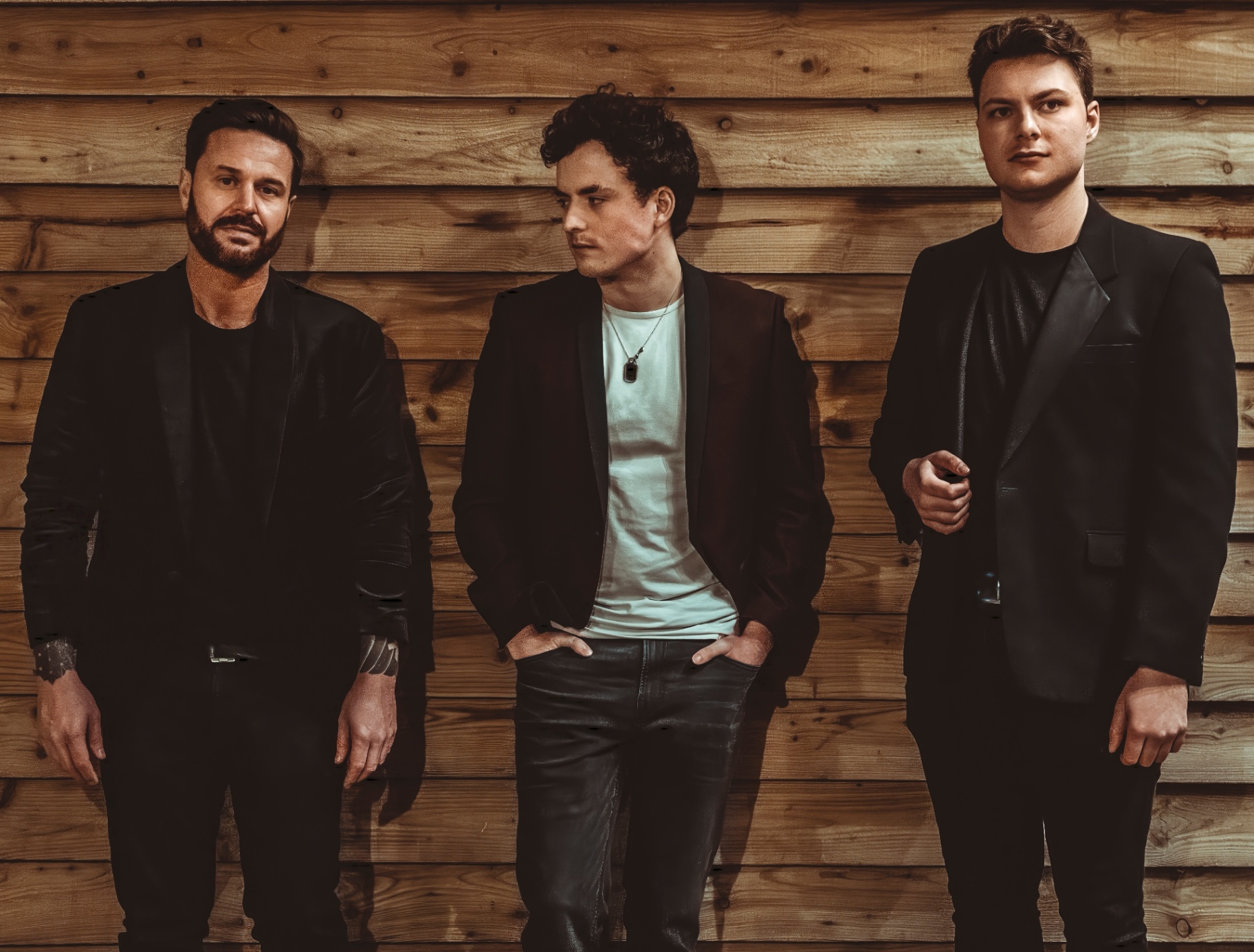 Eleventh Hour: Three band members in dark jackets posing against a wooden wall