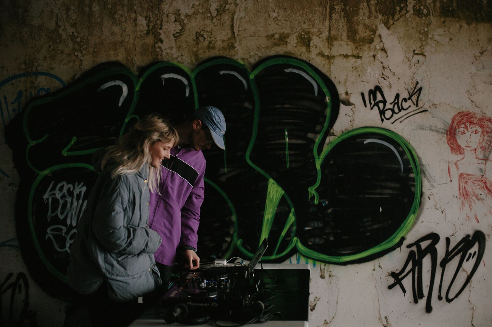 Woman wearing grey coat and man wearing purple coat stand in a tunnel graffiti covered tunnel, over a table with cdjs and laptop on it. In the background there is a piece of graffiti in black and green.