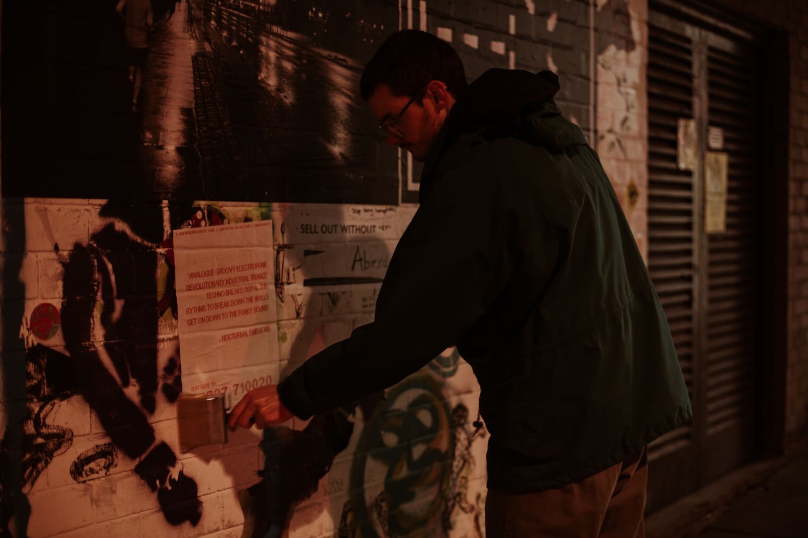Man with wierd and large paint brush and green jacket pasting up poster on white brick wall