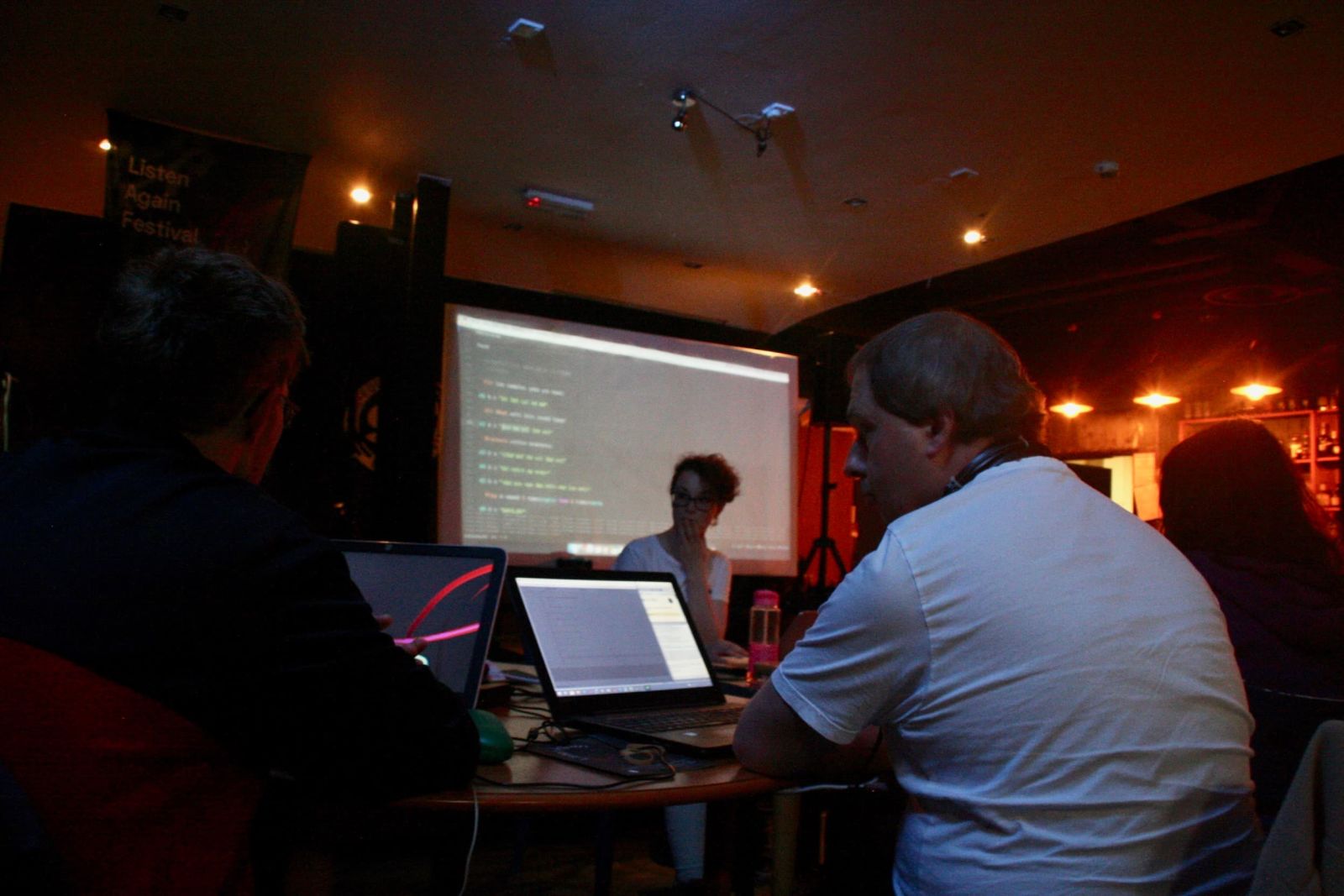 Two men sit at laptops in the foreground whilst a woman in the background is holding the workshops in front of a project screen with code on it