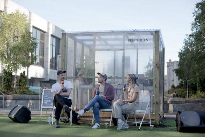 A man and two woman sit on chairs in a semi-circle in front of a greenhouse talking into micorphones