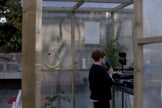 A DJ stands inside a greenhouse talking on a microphone amongst tomato plants and a circular green courgette