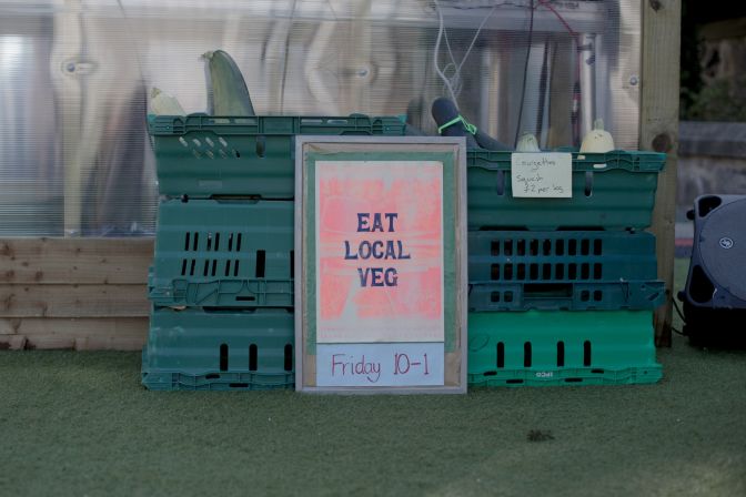 Riso print with words reading eat local veg in bold typography sits in front of plastic boxes containing local fruit and veg.