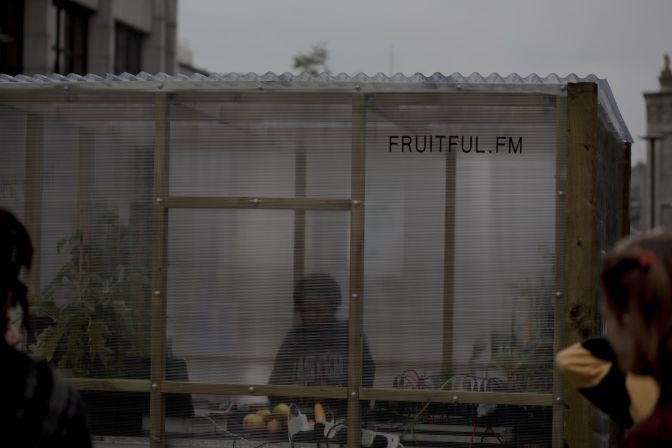 A black woman DJs inside a greenhouse blurred by paneling covering it as two people dance outside