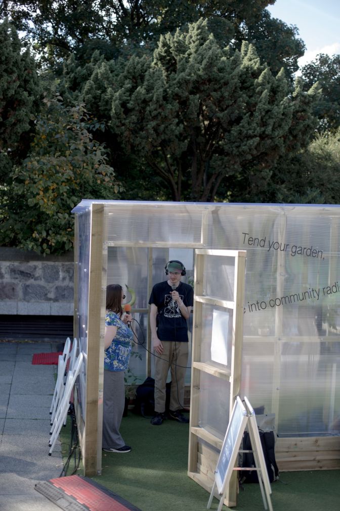 A woman and man stand inside a semi-transparent greenhouse talking on handheld microphones