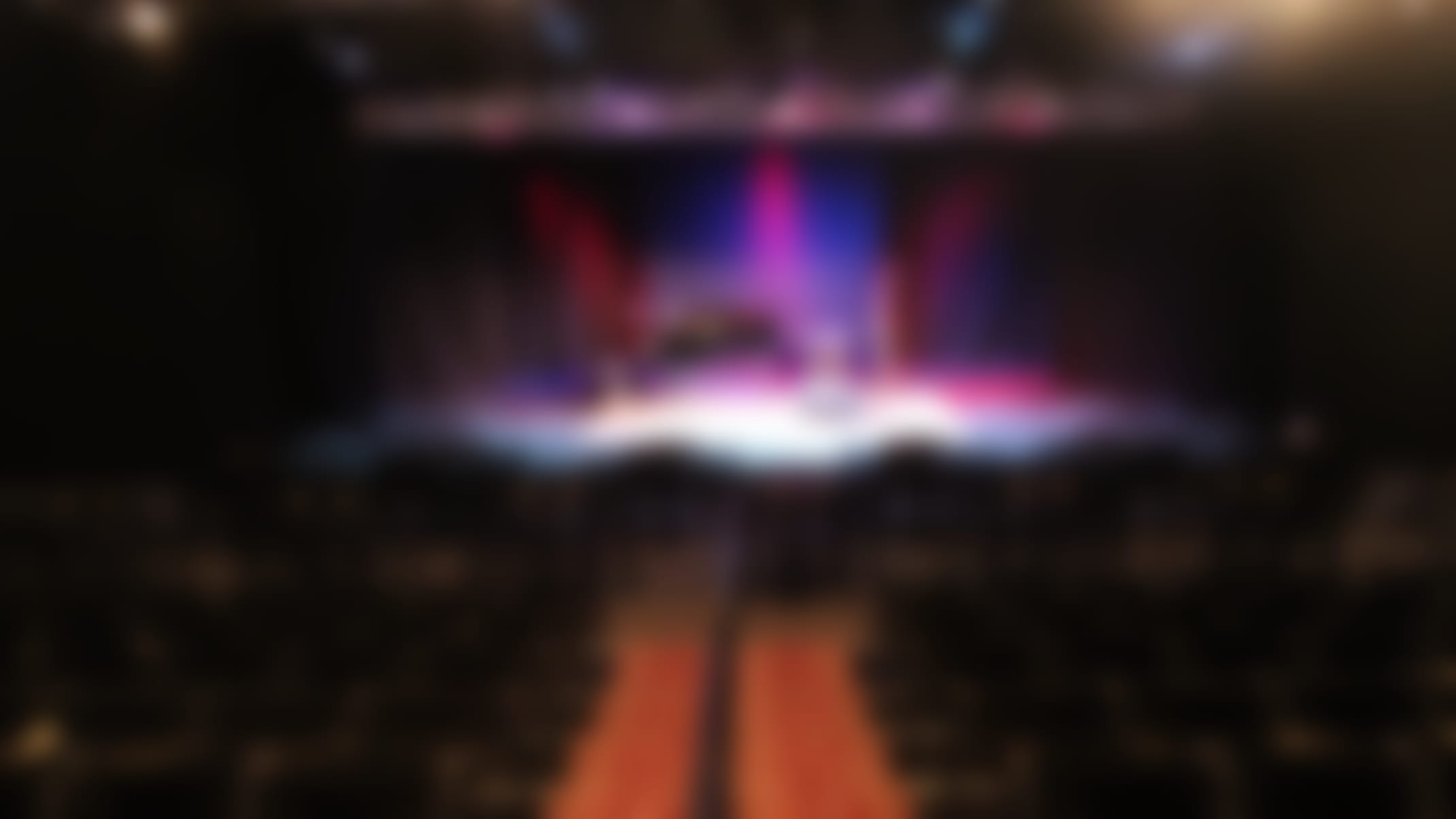 The image shows a dimly lit theater stage with a grand piano in the center, surrounded by rows of empty seats and a red carpet leading up to the stage. The stage is illuminated by colorful lighting, creating a dramatic and atmospheric setting.