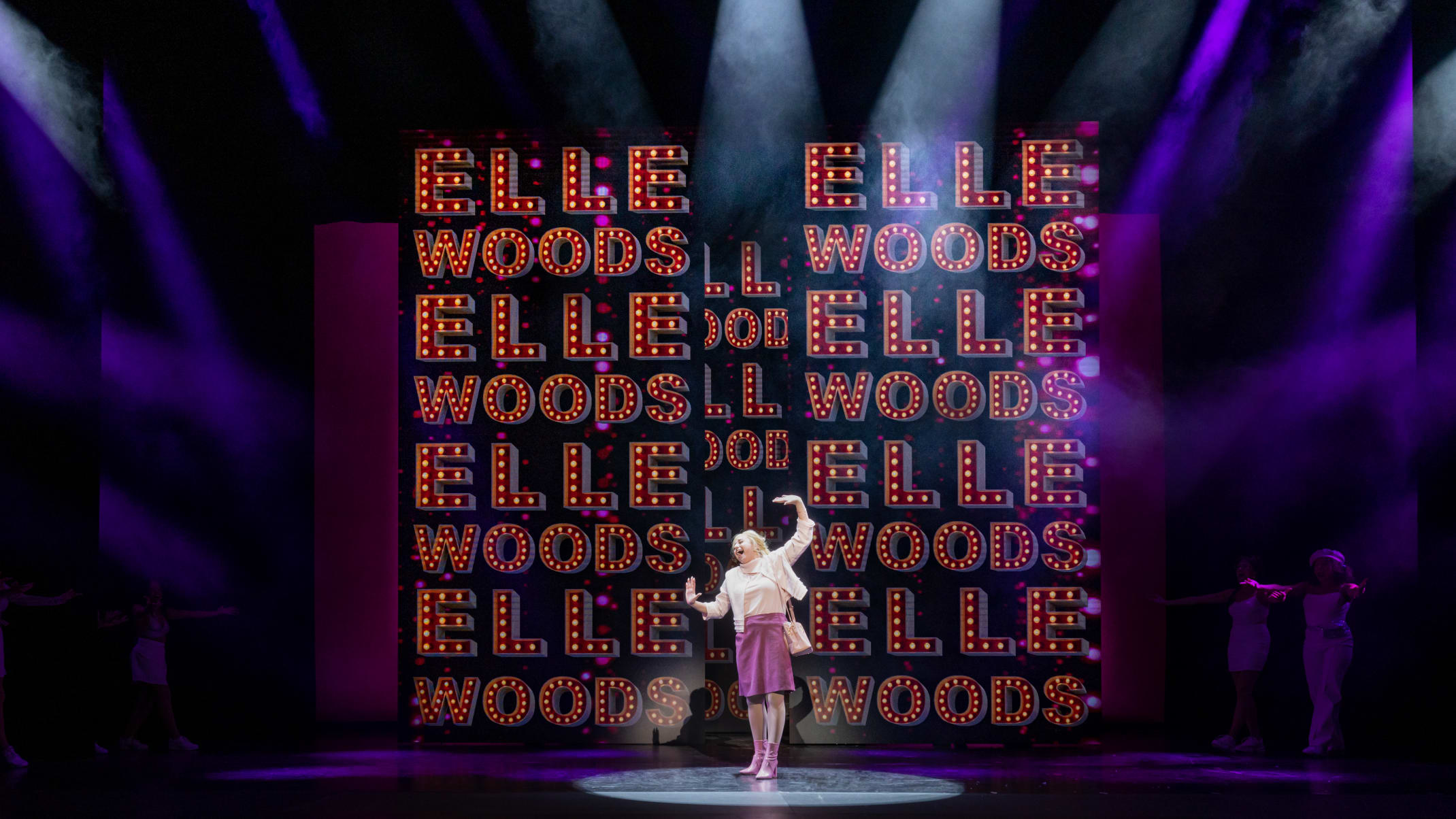 A blonde woman (Elle) standing and posed in front of a wall that shows text saying "Elle Woods" several times.
