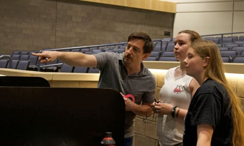 The image shows three people, two women and one man, standing in what appears to be a lecture hall or auditorium. The man is gesturing and pointing, while the two women are listening and observing him. The background features rows of seats and a stage or platform.