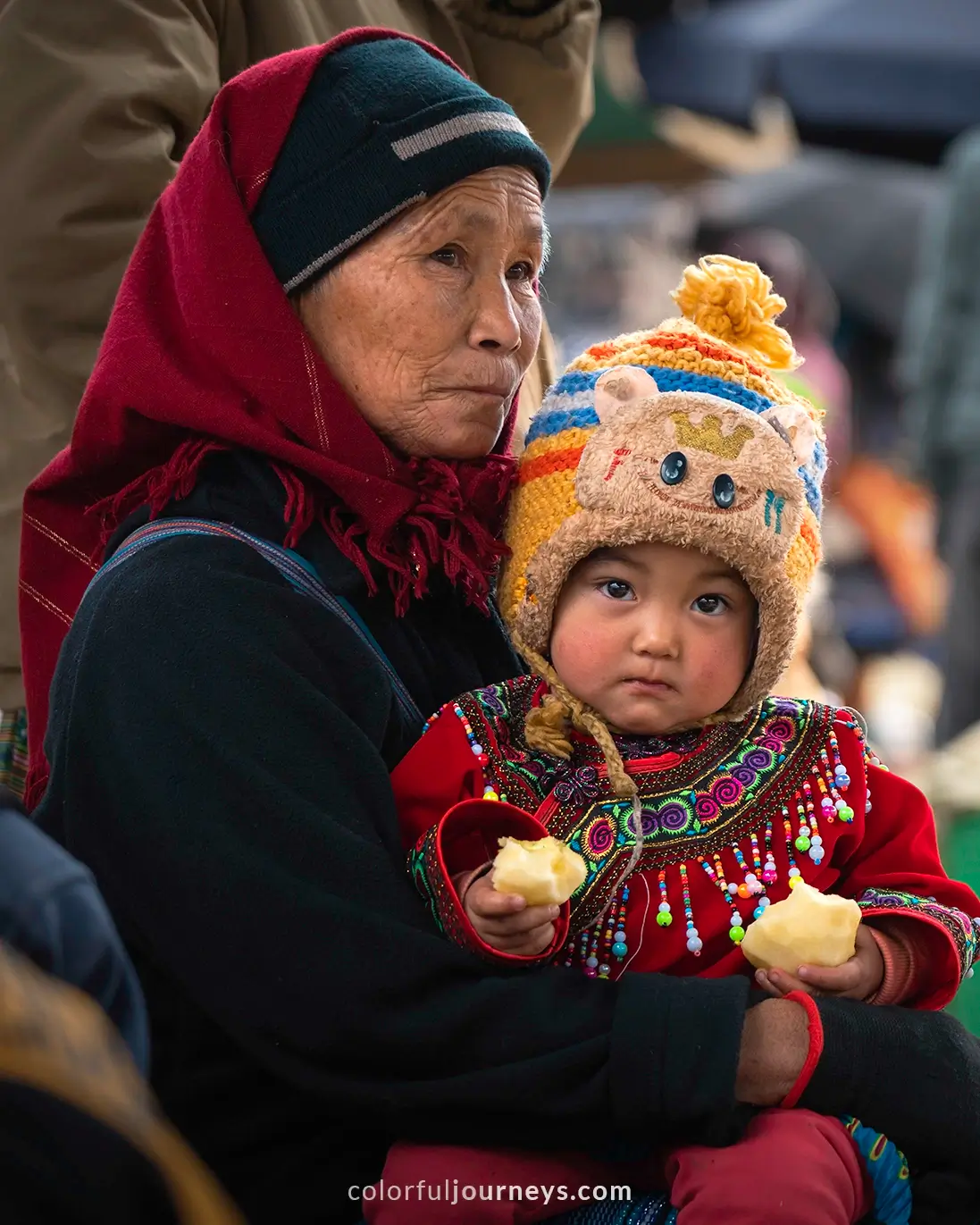 A small girl sits on her grandma's lap while eating an apple at Bac Ha market, Vietnam