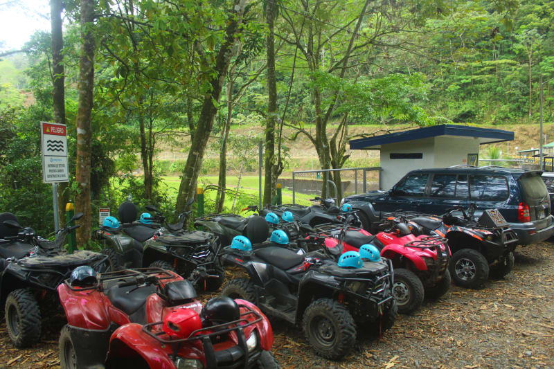 Persona con equipo de seguridad recibiendo instrucciones para manejar un cuadraciclo en un entorno tropical, con más vehículos y vegetación al fondo.