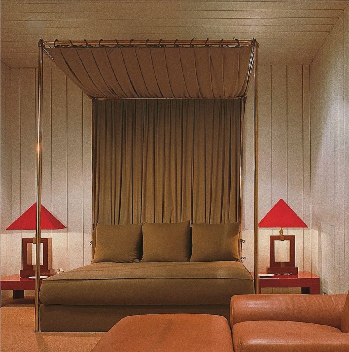 Warm, moody hotel suite featuring a minimalist four-poster daybed with draped canopy fabric, red geometric bedside lamps, caramel leather seating and soft ambient lighting in a retro-inspired luxury i