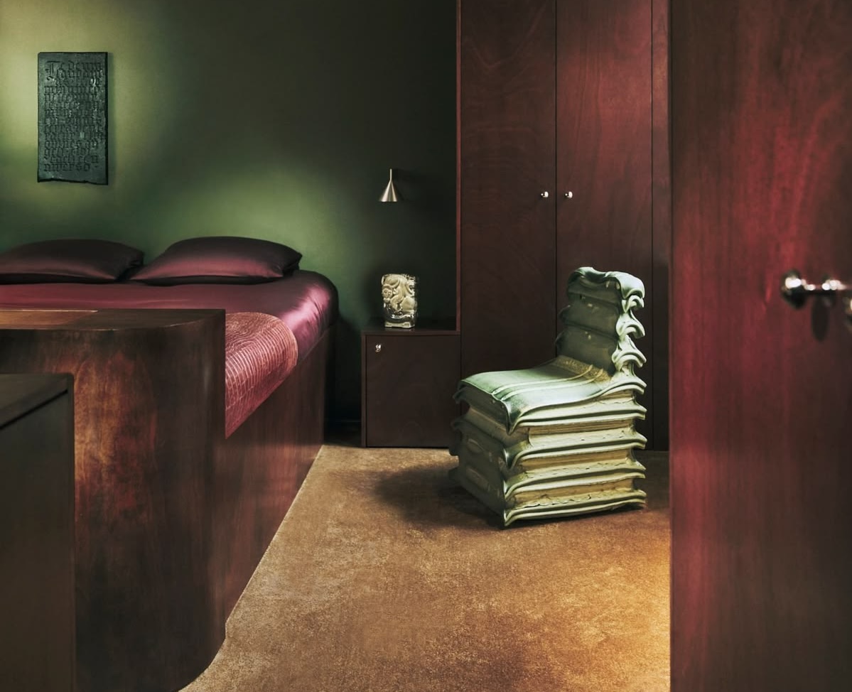 Moody bedroom interior with deep burgundy wood furniture, dark green walls and a sculptural stacked chair centerpiece, styled with rich tonal textures, soft ambient lighting and a minimalist luxury ae
