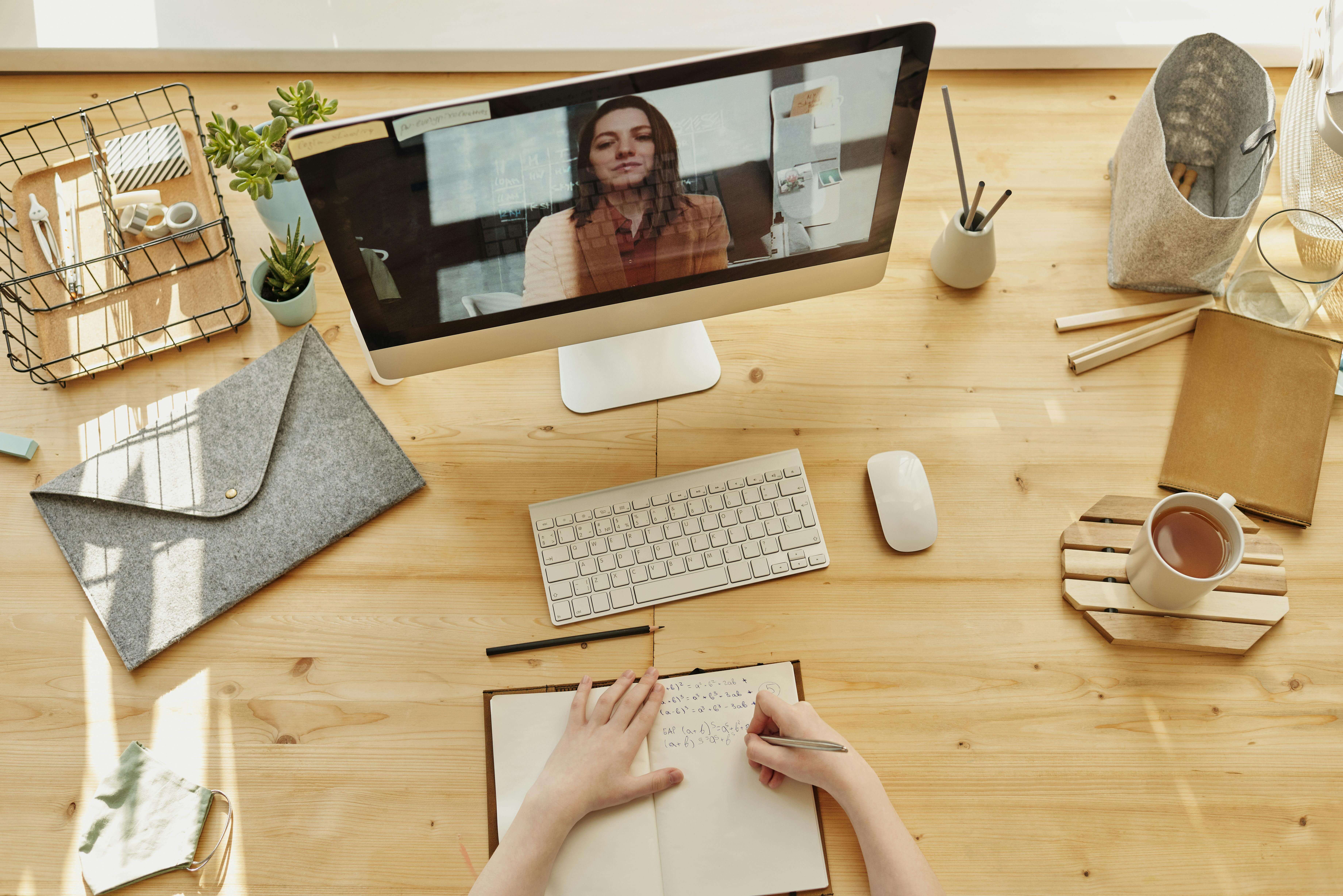 Top‑down view of a tidy desk where someone is taking notes in a notebook during a video call—represents studying and creating cards from lectures.