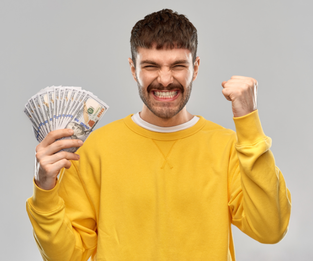 Un hombre feliz con un suéter amarillo, puño en alto, sosteniendo dinero en efectivo. (Aumento de los ingresos como entrenador personal)