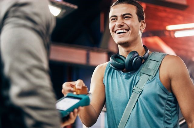  Ein glückliches Mitglied im Fitnessstudio, das eine Transaktion auf einem Mobilgerät abschließt, das ihm von einem Trainer oder einem Mitarbeiter des Fitnessstudios überreicht wird.